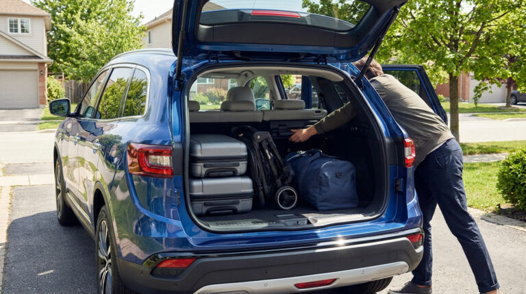 Un homme range des bagages et une poussette dans le coffre ouvert d'un Renault Symbioz bleu stationné dans une allée.