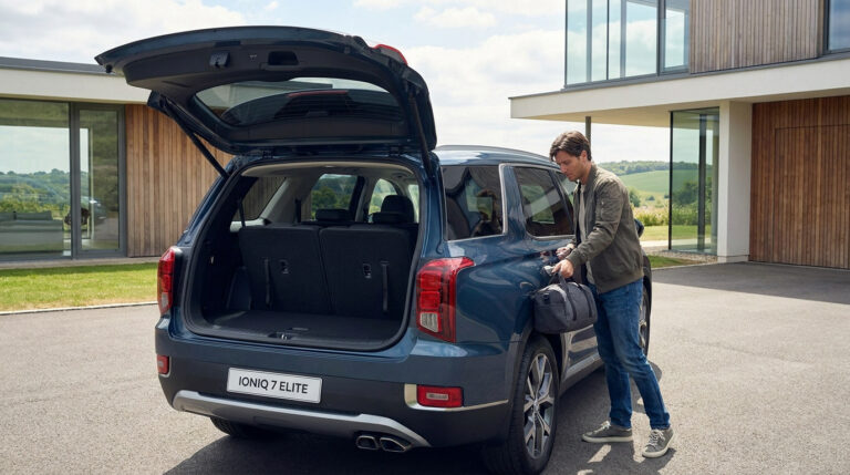 Un homme charge un sac dans le coffre ouvert d'un SUV bleu Hyundai Ioniq 7 Elite stationné devant une maison moderne.